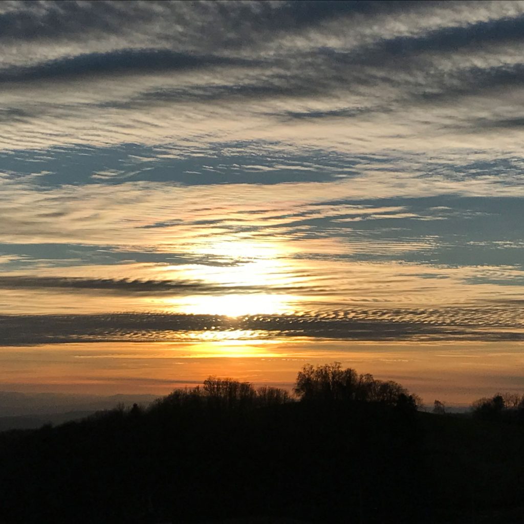 wellige Wolkenstruktur, die die untergehende Sonne bricht, unten Hügel mit noch unbelaubten Bäumen, alles im Gegenlicht